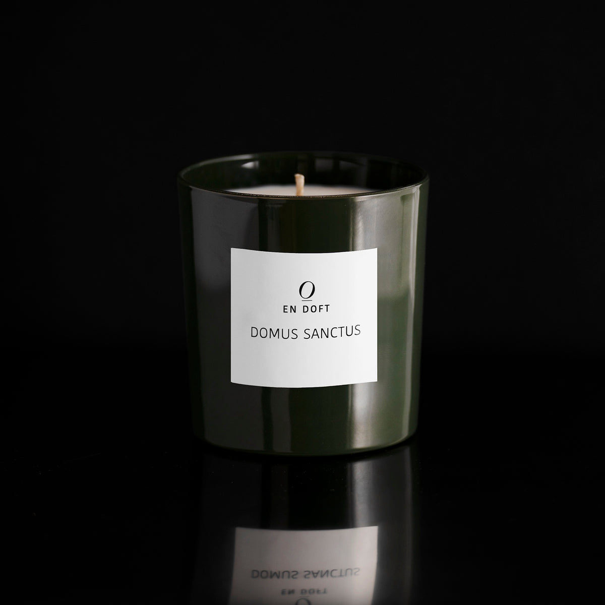 DOMUS SANCTUS scented candle by En Doft in a dark green glass vessel with a white label, on a black reflective surface.