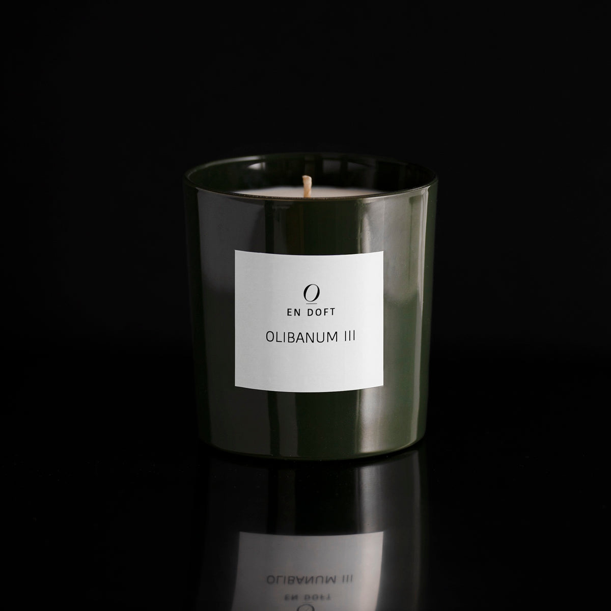 OLIBANUM III scented candle by En Doft in a dark green glass vessel with a white label, on a black reflective surface.